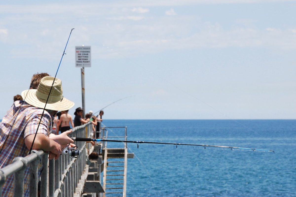 shutterstock_1416000098 | Island Getaways People fishing from a seaside pier, facing the water under a blue sky with the ocean in view.
